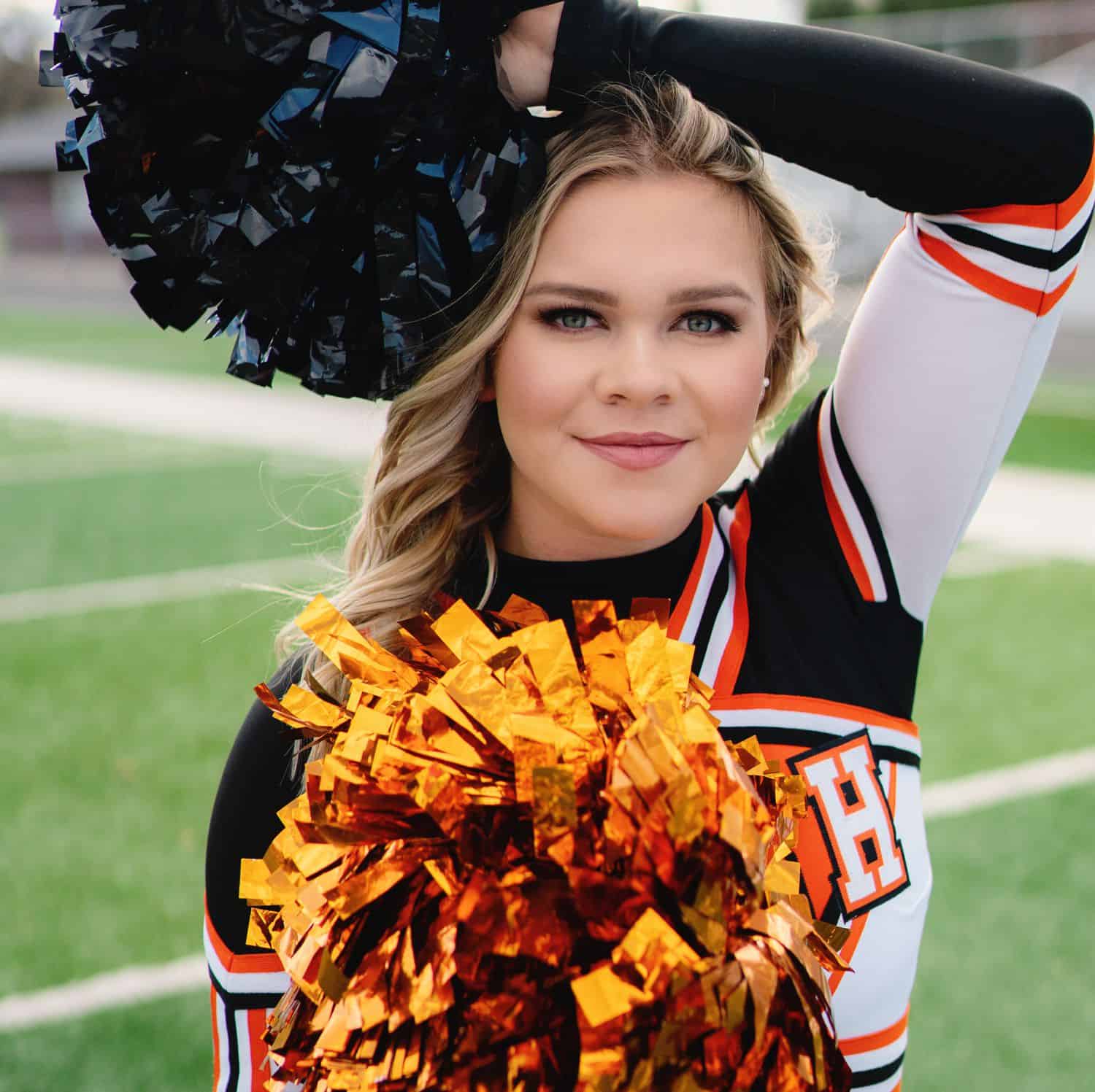 A senior girl cheerleader stands on a football field with her pop-poms on display