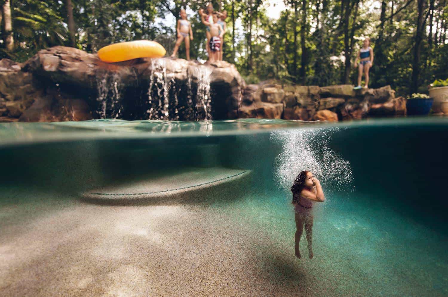A split-level underwater photographs depicts an above-grown human-made waterfall spilling into a swimming pool where a little child swims toward the surface while holding their nose.