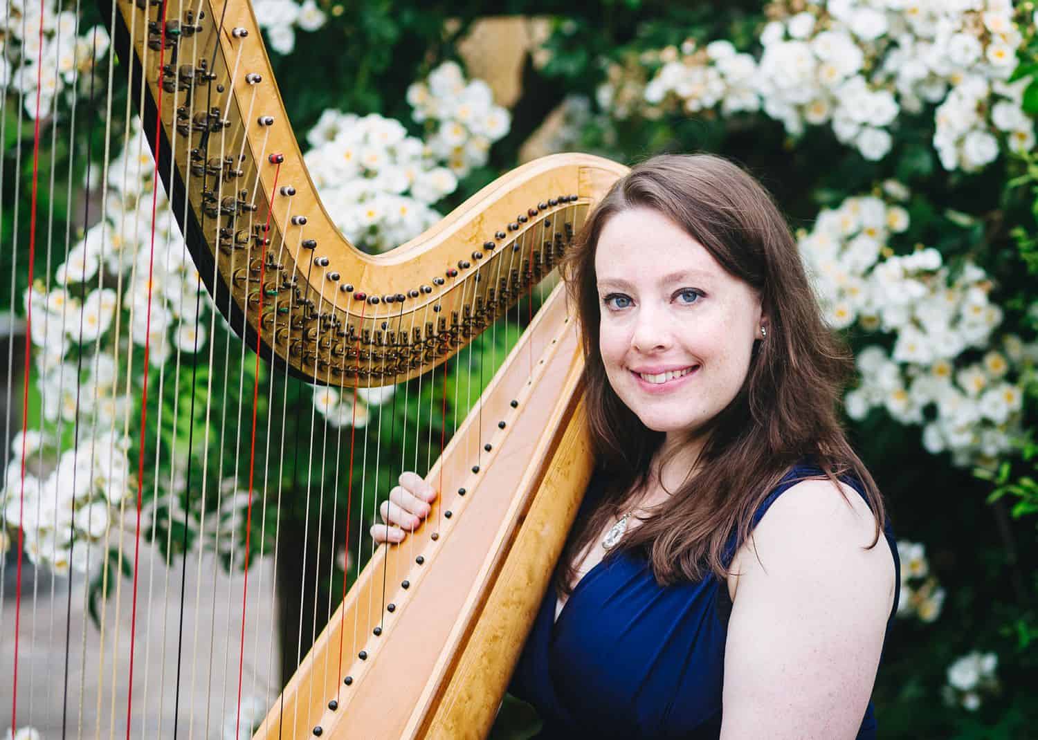 A brunette woman in a blue dress sits in front of a flowering bush with her harp