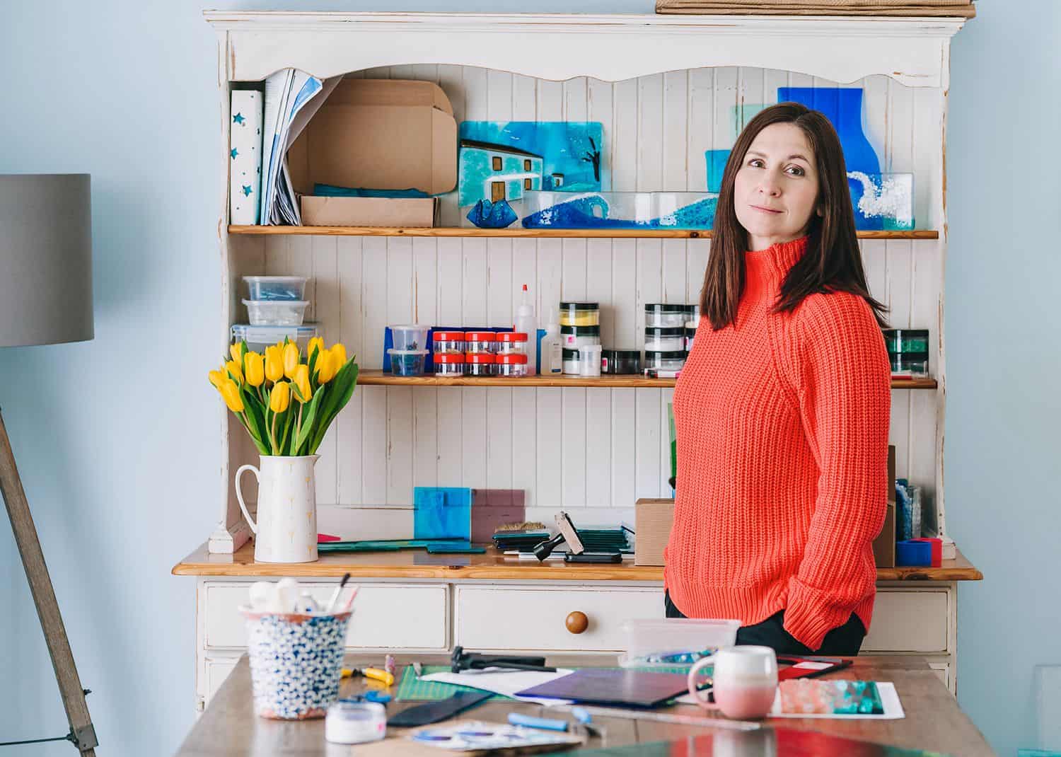 A brunette woman in a bright orange sweater stands in her shop surrounded by artwork