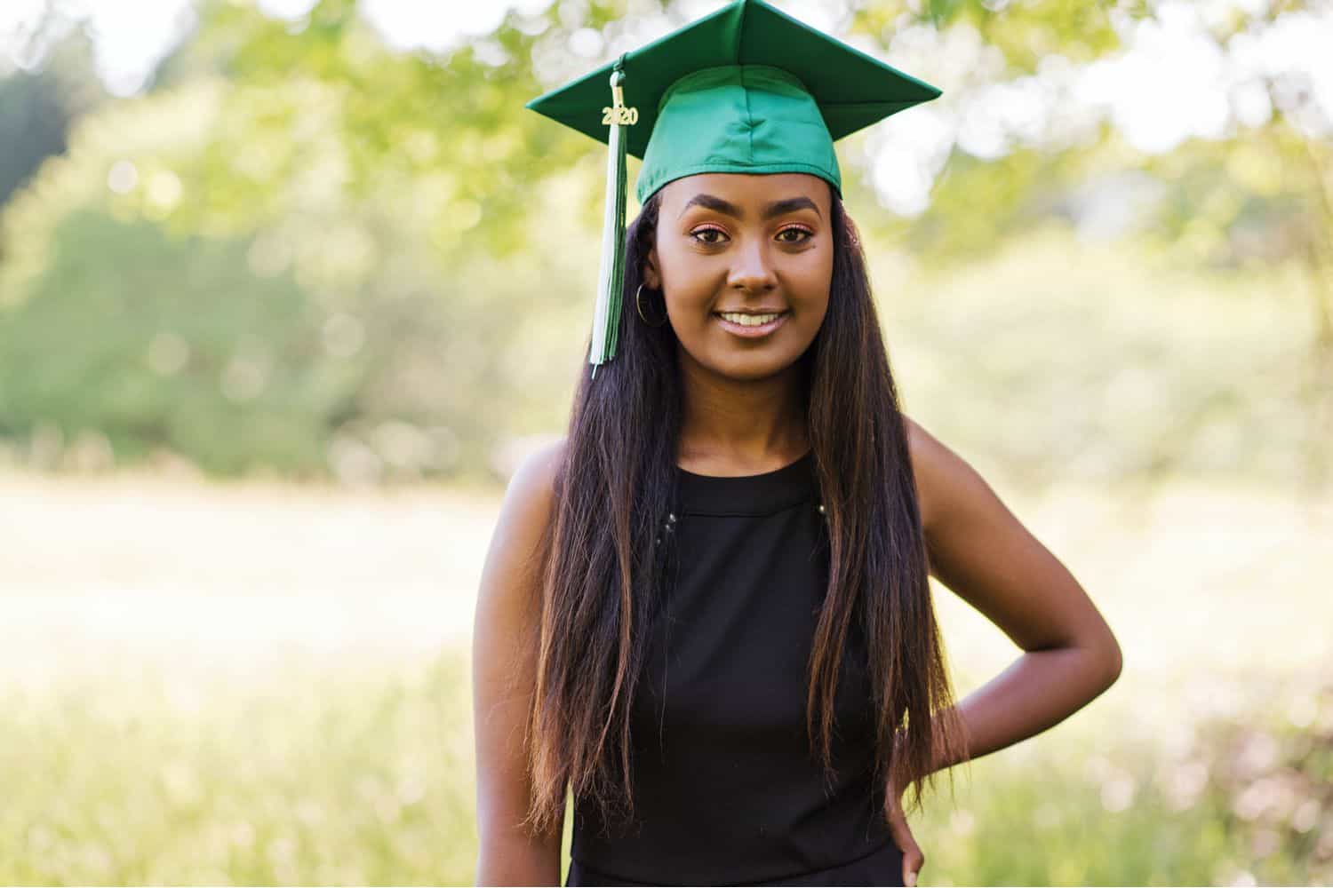 A high school senior girl poses in a field wearing a black dress and a green graduation cap