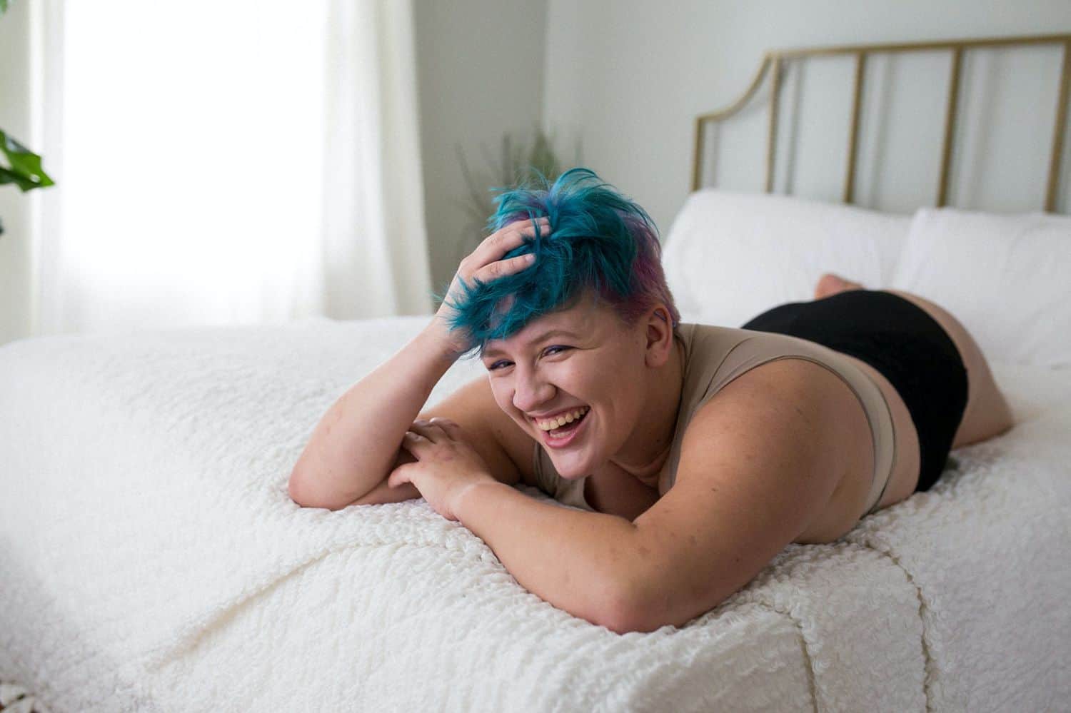 A person with fair skin and short blue hair lies on their tummy on a white bed with their head in their hand. The model is laughing as they are photographed by boudoir photographer Kinzie Ferguson, the Empowerment Photographer