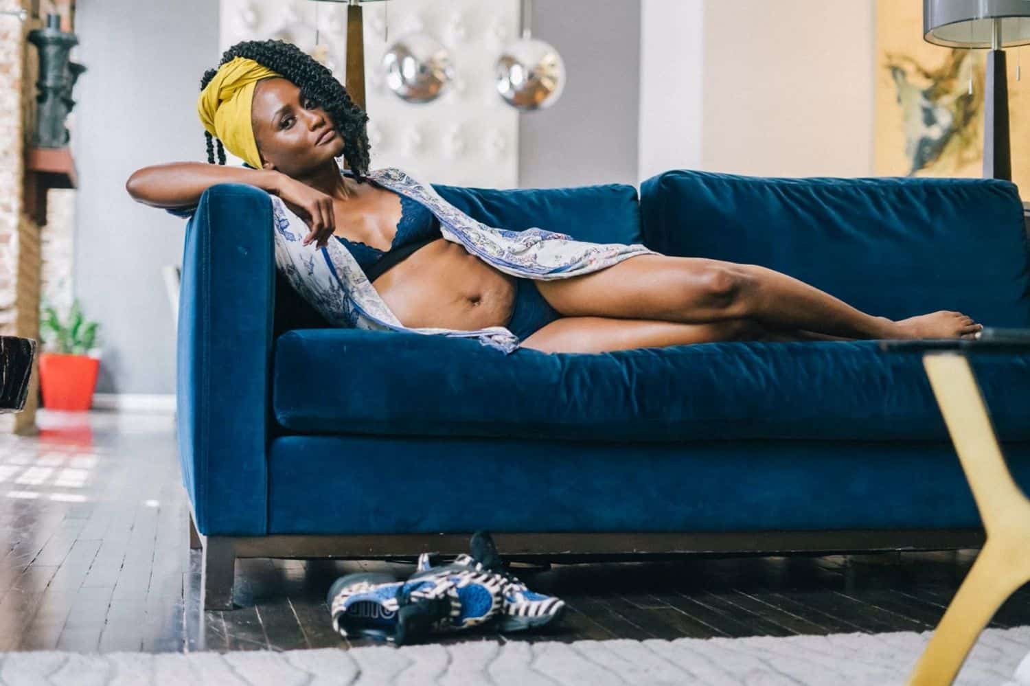 A person with dark skin wearing blue lingerie reclines on a blue velvet sofa. They wear a bright yellow head wrap and look serenely at the camera as they are photographed by Toni Black