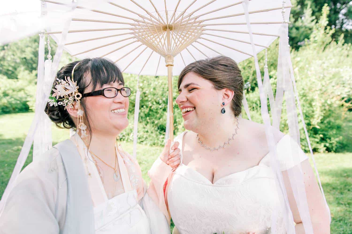 Two brides stand under a paper umbrella laughing