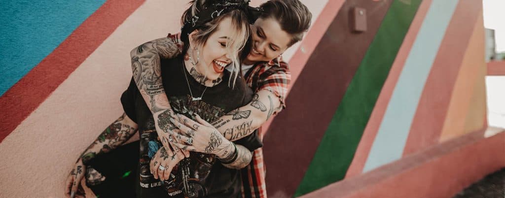 Two people with tons of beautiful tattoos snuggle close with their arms around each other. They are both laughing. The wall behind them is painted like a vintage rainbow.