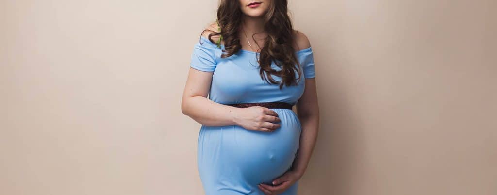 A pregnant person stands in front of a beige studio backdrop with her hands cradling her belly. She is wearing a pale blue, off-the-shoulder dress, through which her belly button is barely visible. The mom-to-be's long brown hair curls gently over her collarbone.