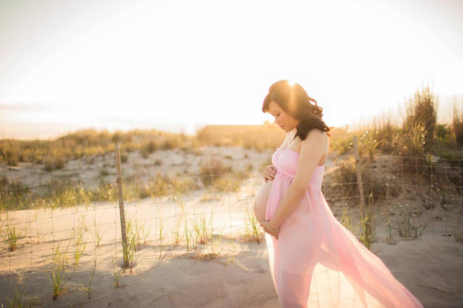 A pregnant woman in a flowing, pink, semi-sheer gown cradles her belly and gazes down toward her unborn child. She's standing on a beach shortly before sunset with dune grass and a bright sky behind her.