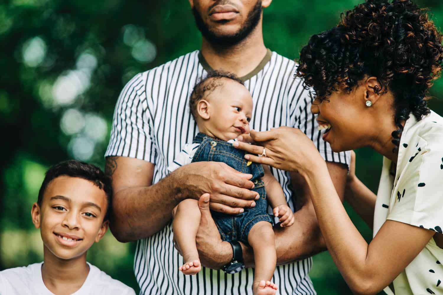 A Black family stands close together for an outdoor portrait. The father is holding their infant baby, while the mother tickles the baby's cheek. An older son stands close to his dad, smiling.