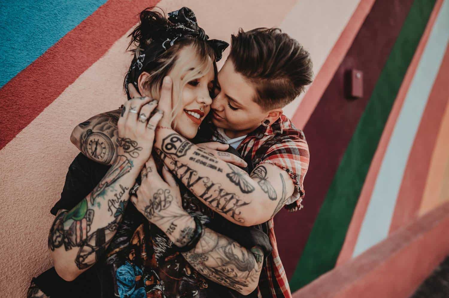 A heavily tattooed couple holds one another close while standing in front of a rainbow-painted wall.