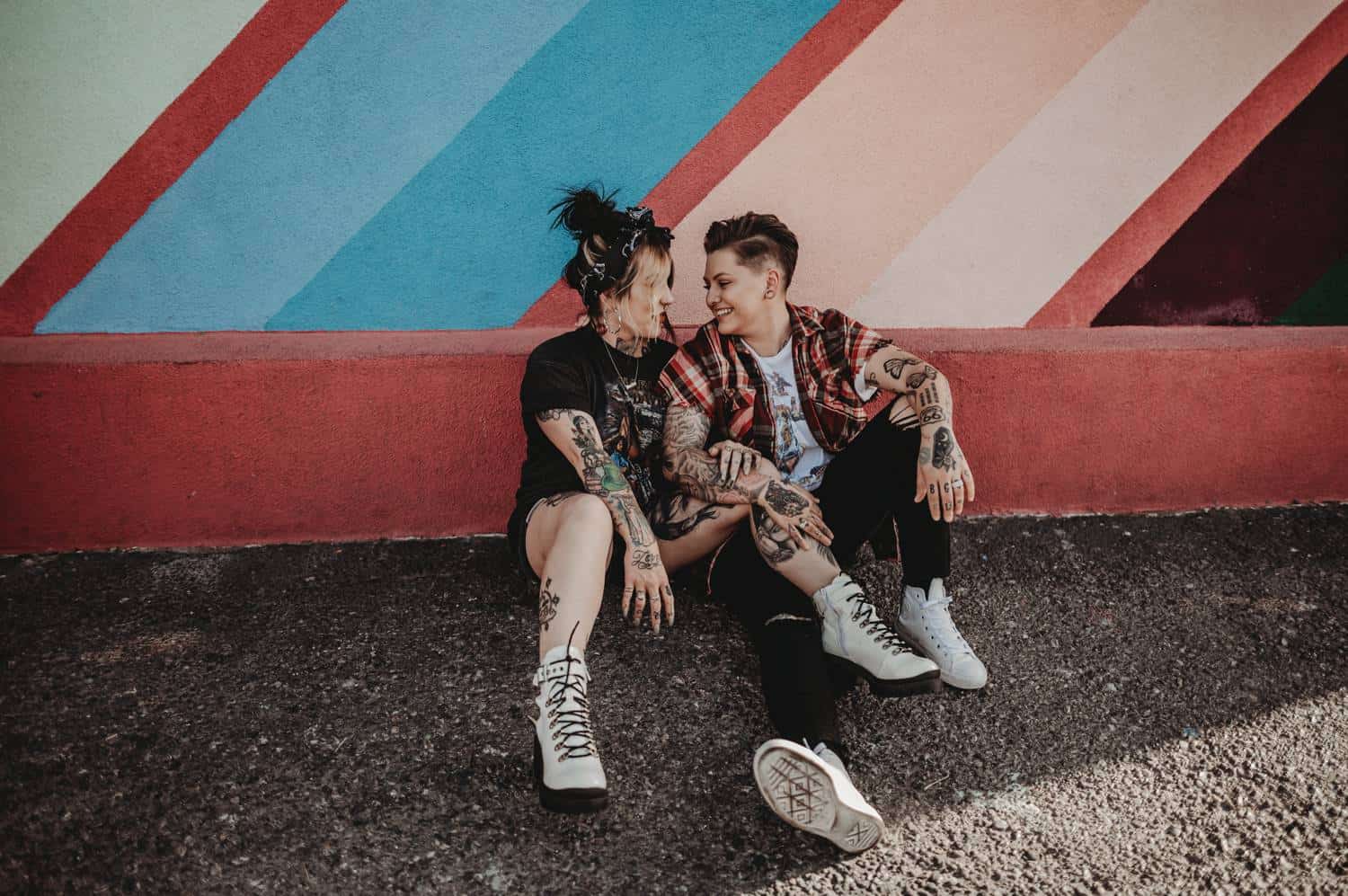 A couple sits on the asphalt and lean against a rainbow-painted wall. They are both casually dressed and covered in bold tattoos. They are both smiling and looking at each other lovingly.