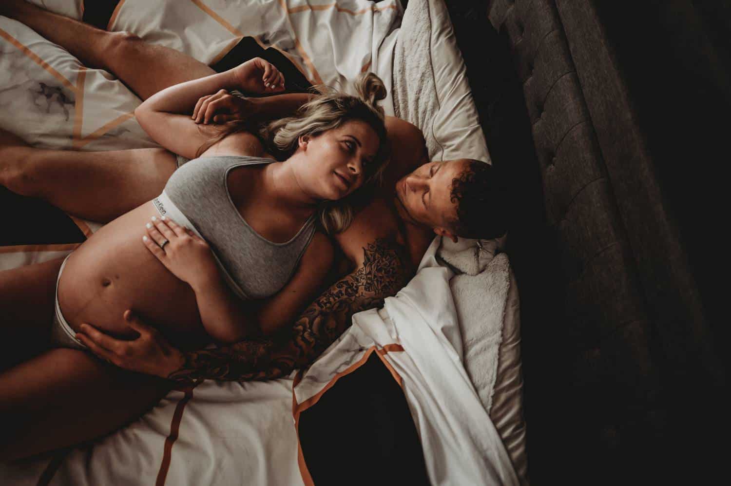 A couple lies on their bed in a dimly lit room. They are photographed from above, with the camera looking down on them. The woman lounged against her partner's chest, with her hand draped across her pregnant belly. She's wearing a gray Calvin Klein underwear set, and her partner's tattooed arms are wrapped around her as they look at one another.