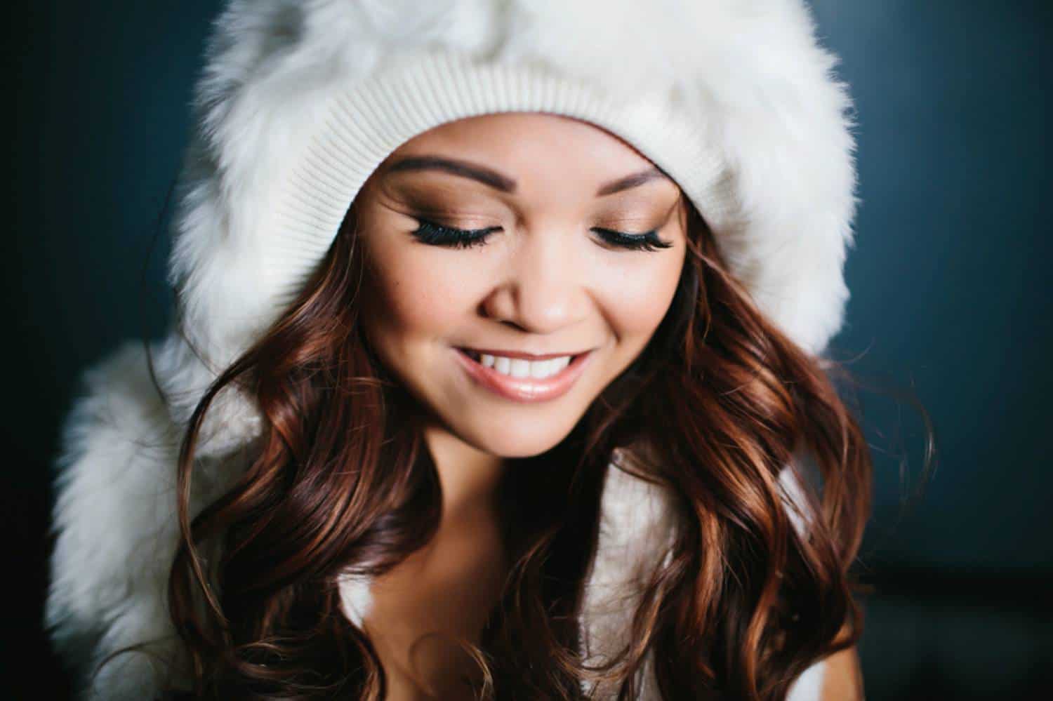 A woman with wavy brunette hair look down while wearing a furry white hood.