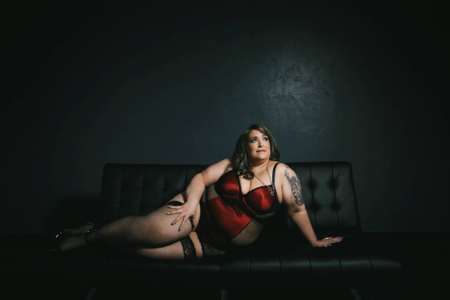 A woman in a red negligee and thigh-high tights reclines on a tufted leather sofa in a dark photography studio.