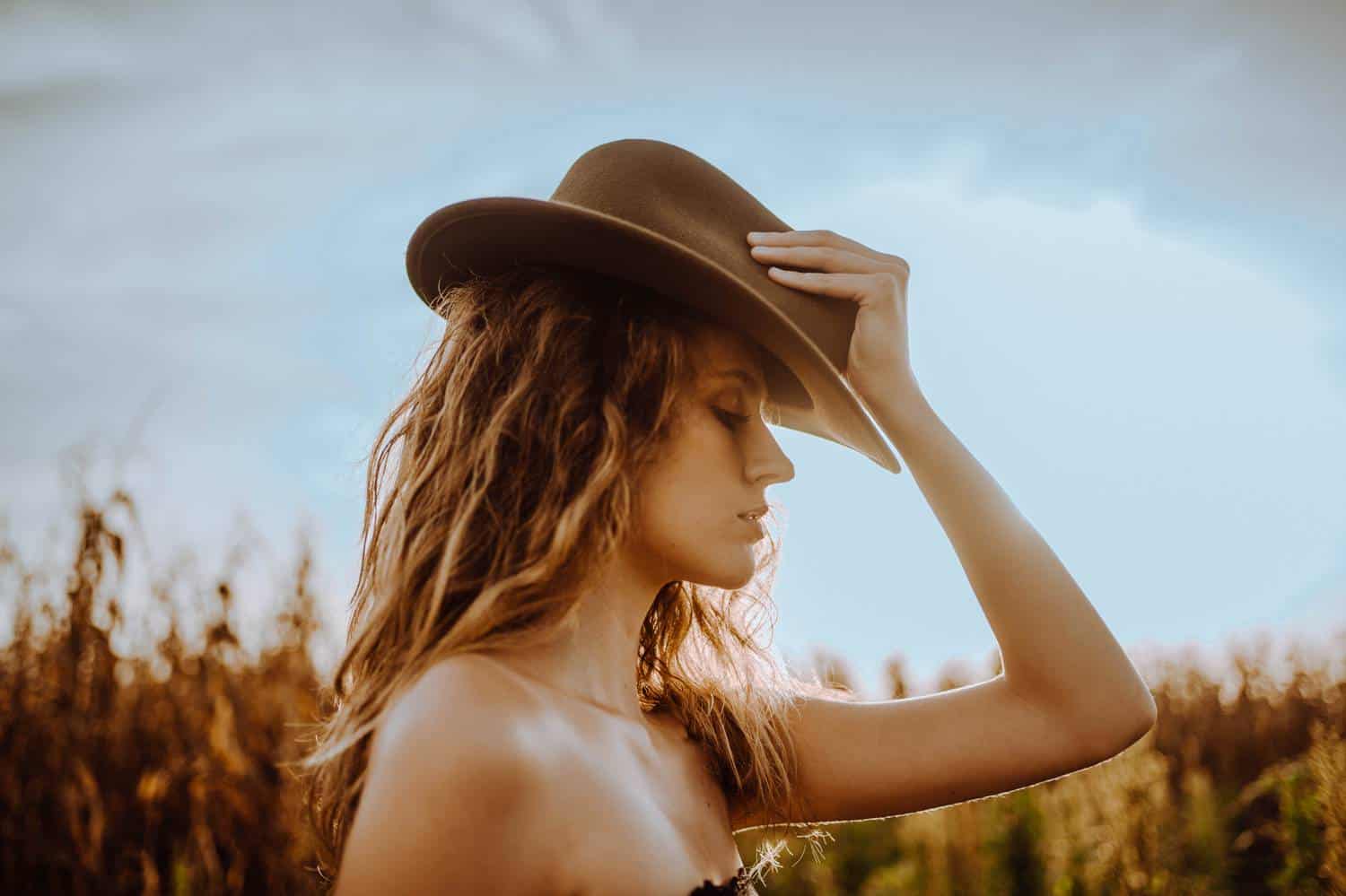A woman with bare shoulders and blonde hair stands with her profile to the camera, adjusting her felt cowboy hat in the middle of a field.