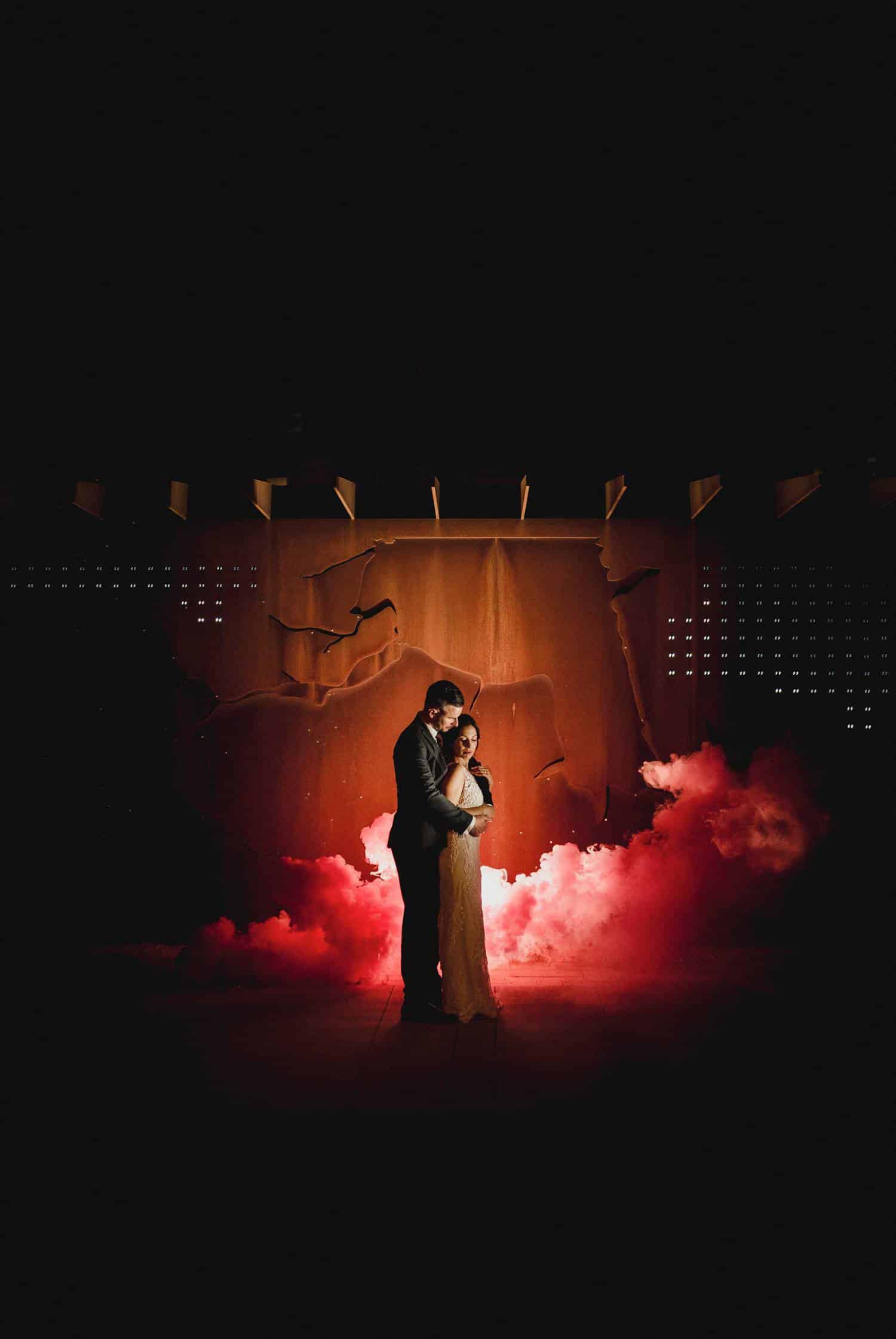 A modern, angular background is emphasized by red smoke that filters behind a bride and groom, who stand back-to-front looking toward the ground. Black & Gold Photography are known for making immaculate wedding portraits using off-camera flash.