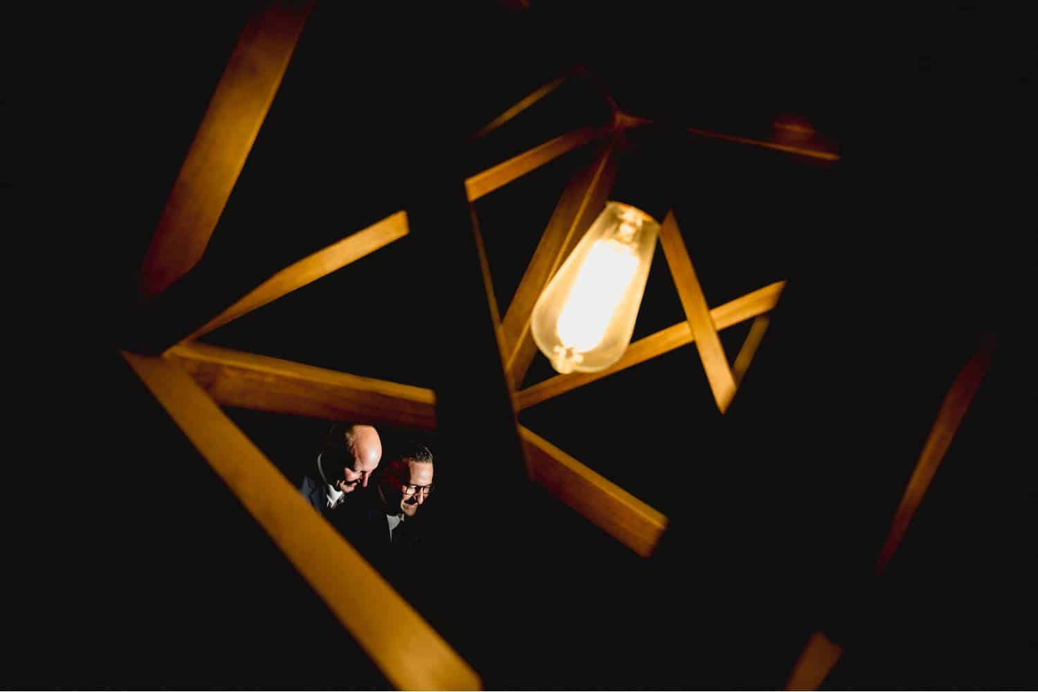 Two grooms' faces are highlighted between the angular bars of a modern light fixture. Black & Gold Photography made this unique wedding portrait using off-camera flash.