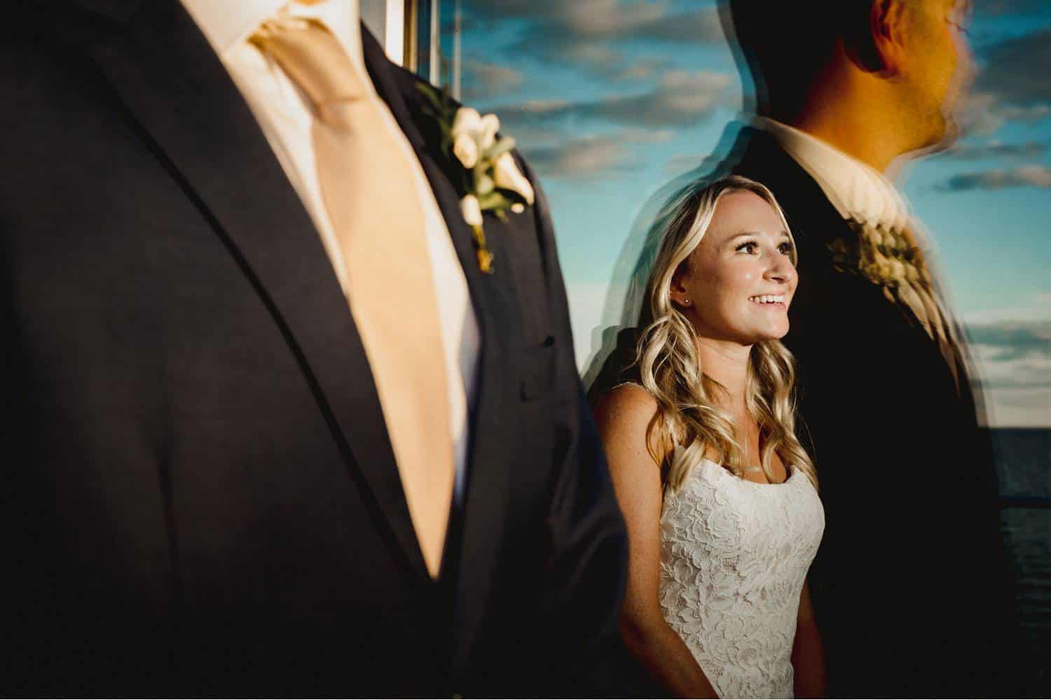 A bride stands indoors facing a flash with a smile on her face. The groom stands closer to the camera, on the outdoor side of the window through which you can see the bride. His form and the blue sky are both reflected off the glass, surrounding the bride. Black & Gold Photography creates wedding portraits using off-camera flash.