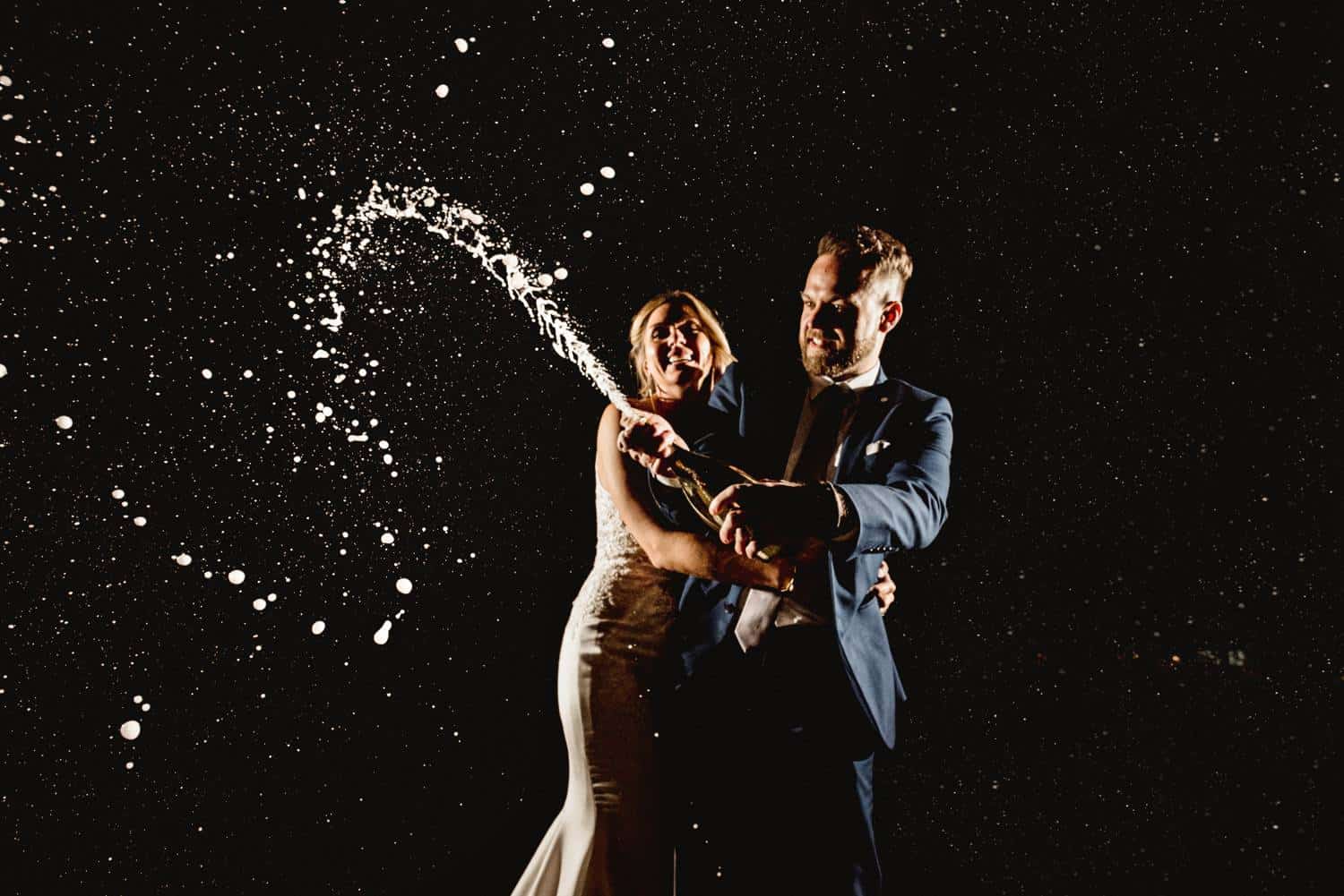 A wedding couple stands against a nighttime backdrop and pops a fresh bottle of champagne. The bride's face is jubilant and her arms encircle the groom as he pops the bottle's cork with determination on his face. Black & Gold Photography makes amazing wedding portraits using off-camera flash.