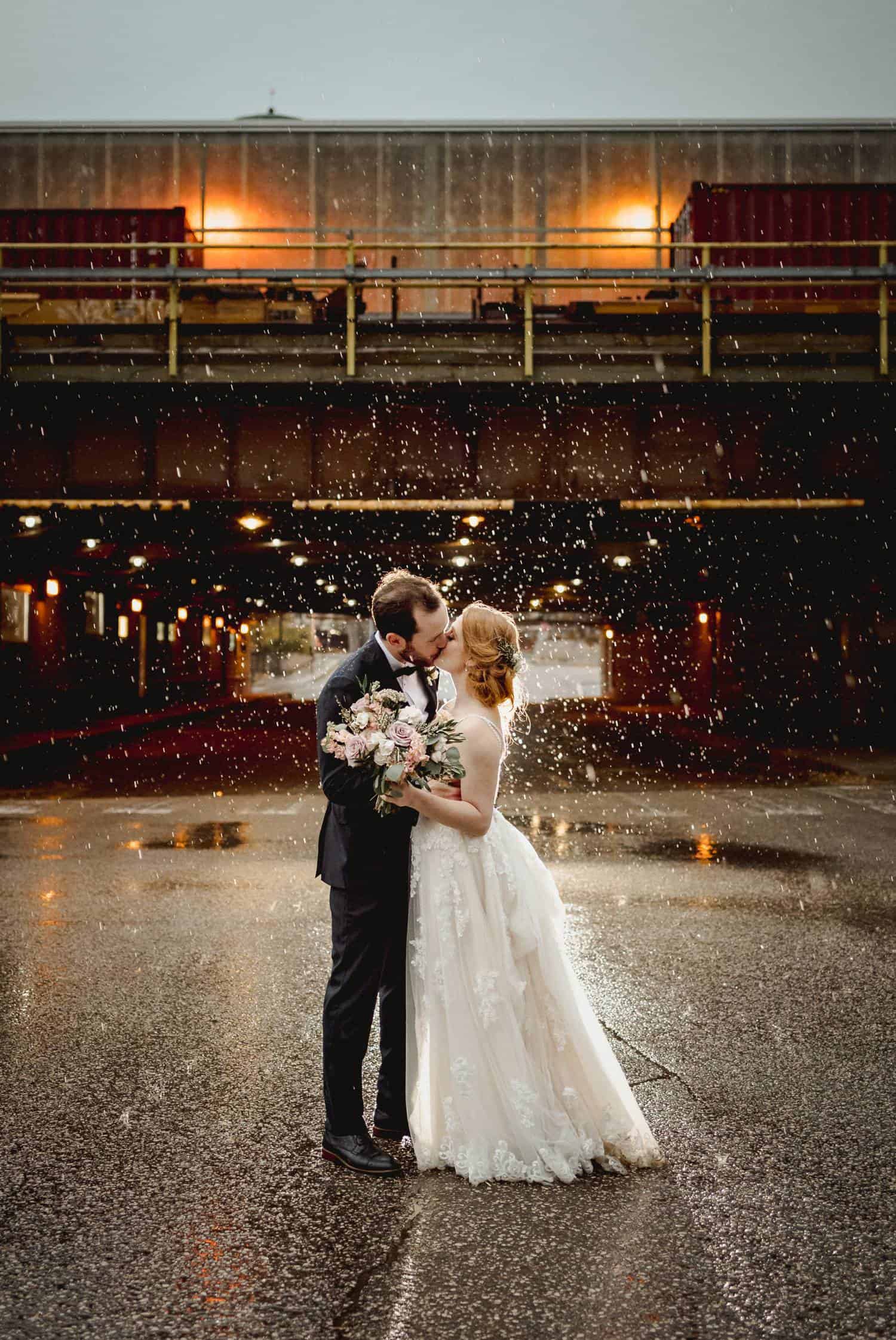 A bride and groom stand kissing in the middle of the road. A train trestle looms behind them, and rain pours all around them. Black & Gold Photography uses this photograph to teach photographers how to make wedding portraits using off-camera flash.