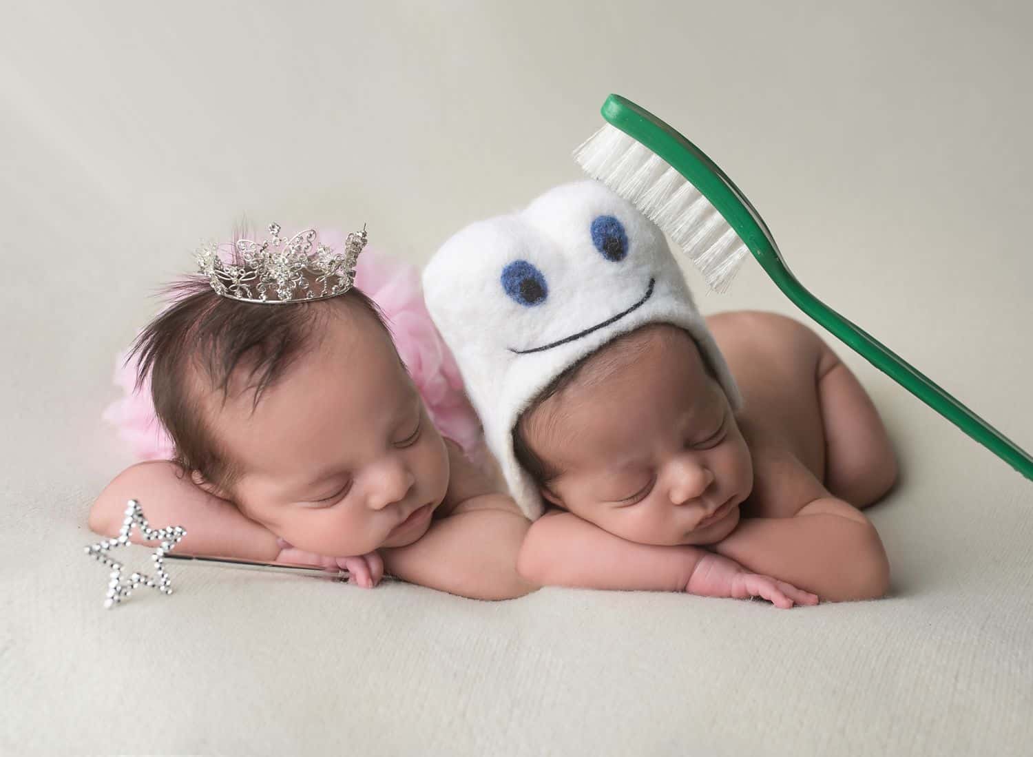 The best newborn photography props include a tiny tooth hat paired with an oversized green toothbrush, shown here on a baby boy. His twin sister wears a pink tutu and tiny tiara, and she holds a glittering wand in her hand as she sleeps.