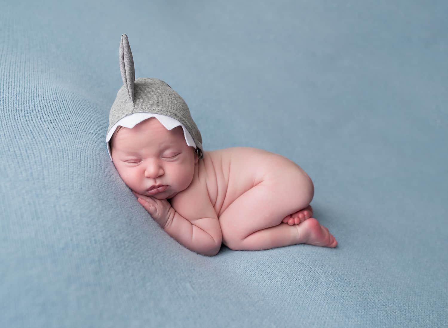 The best newborn photography props don't have to be complicated. This simple setup shows a newborn in froggy pose asleep on a blue cushion while wearing a tiny cotton shark fin hat.