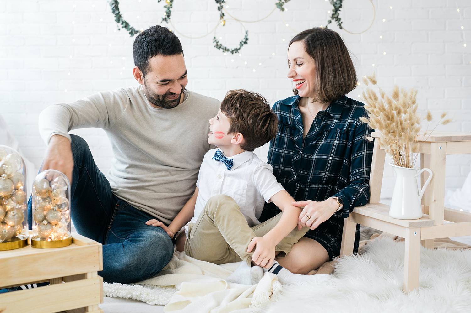 In this photo by Bruna Rico, a dad, mom, and young son sit on a white faux fur in a white room. Ropes of garland are hung behind them, and small wooden crates sit beside them holding gold ornaments. Christmas Mini Session: Ideas Families will ADORE!
