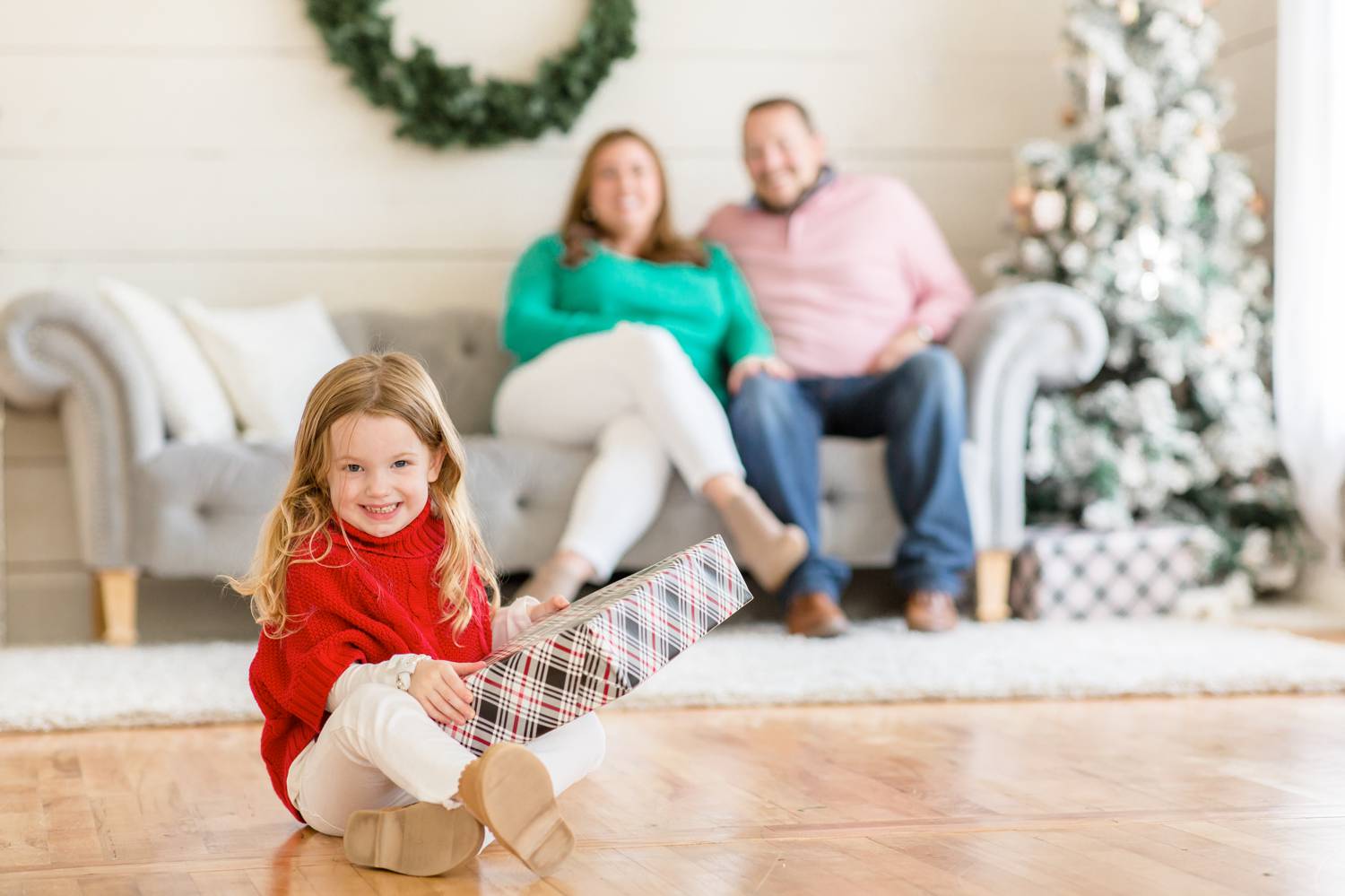 In the photo by Rebecca Rice, two parents sit on a silver sofa, blurry in the background. In the foreground and in focus, a little girls sits on the ground holding a wrapped present as she smiles at the camera. Christmas Mini Session: Ideas Families will ADORE!