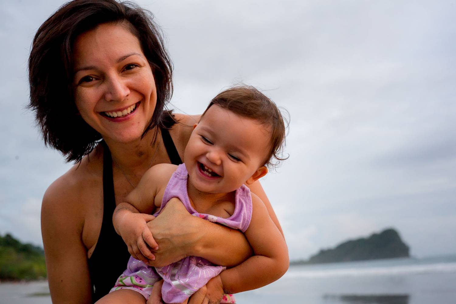A photo by Kevin Heslin depicts a mom smiling with her laughing baby in her arms as they play on a beach.