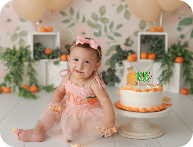 A baby girl in a peach-colored dress with "one" embroidered on the front sits next to a white cake with orange frosting and a "one" topper, surrounded by a floral backdrop, greenery, and oranges. The baby has cake on her face and hands, indicating a "smash cake" first birthday celebration.