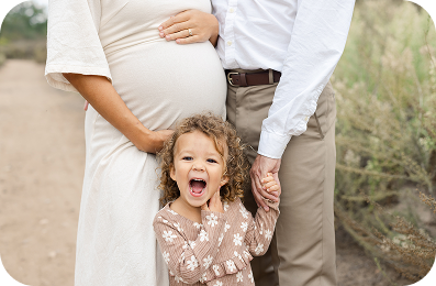 A pregnant woman in a cream dress holds her belly while standing next to a man in a white shirt and khaki pants, and their young daughter with curly hair and a floral shirt stands in front, smiling with her mouth open and holding their hands. The family is outdoors with a dirt path and foliage in the background.