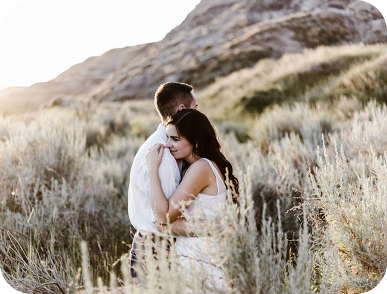 A young couple embraces in a field of tall grass, with a hill in the background. The woman, wearing a white dress, wraps her arms around the man, who is wearing a white shirt.
