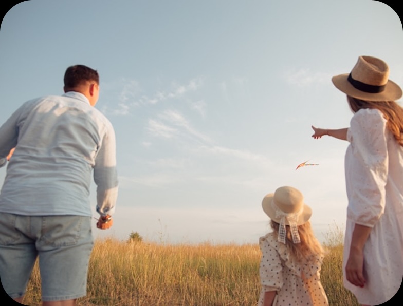 A family of three is flying a kite in a field of tall grass under a blue sky. The mother, wearing a hat and white dress, points to the kite, while the father holds the string and the daughter looks up at the kite, also wearing a hat.