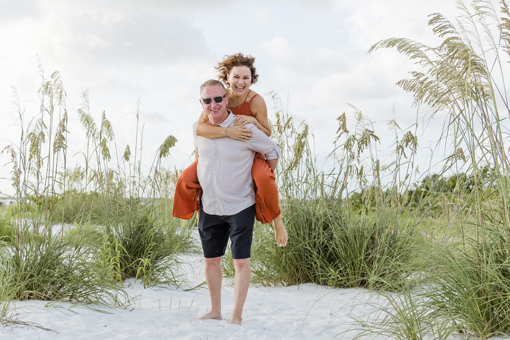 A smiling man in sunglasses gives a piggyback ride to a laughing woman on a sandy beach with tall grasses. The man wears a light-colored shirt and dark shorts, while the woman is in an orange jumpsuit.