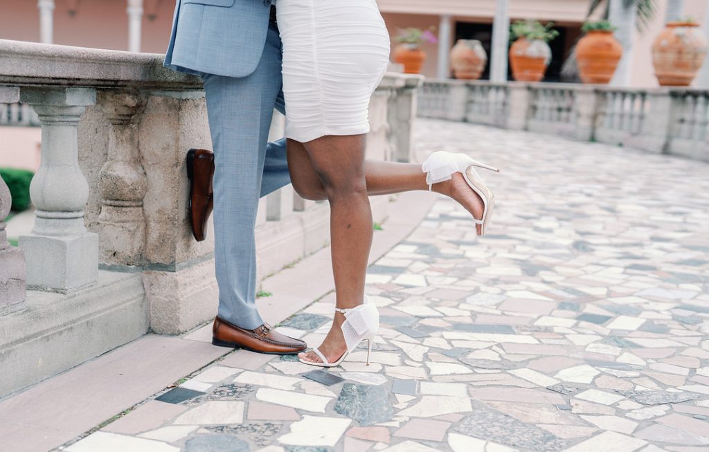 A close-up shot focuses on the legs of a couple, where a woman in a white dress and white bow-accented heels playfully lifts one leg while standing close to a man in a light blue suit and brown loafers on a stone-paved terrace.