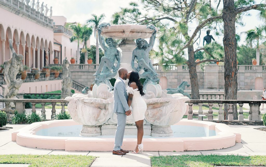 A well-dressed couple kisses while holding hands in front of an ornate, classical stone fountain in a formal garden with statues and palm trees.