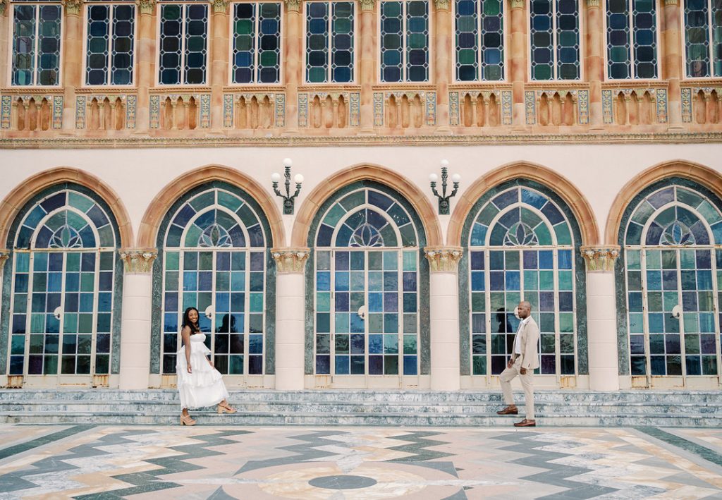 A smiling woman in a white tiered dress and a man in a beige suit stand apart on the steps of an ornate building featuring large, colorful stained-glass arched windows and a patterned stone courtyard.