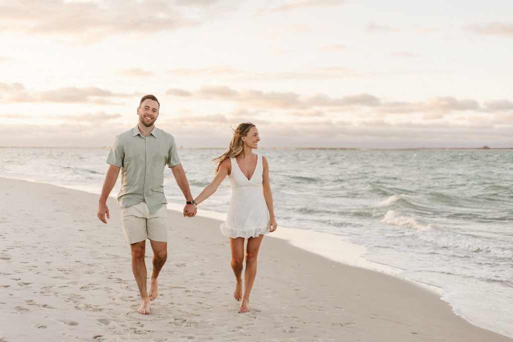 A smiling young couple walks hand-in-hand along a sandy beach shoreline at sunset, with the man in a light green button-down shirt and shorts looking toward the camera while the woman in a white sundress looks at him.