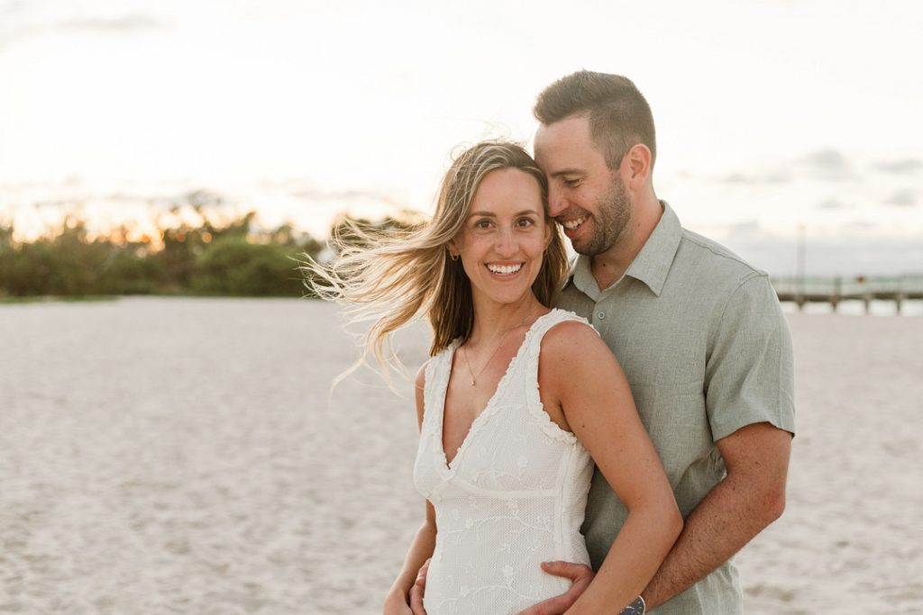 A smiling man in a light green shirt embraces a woman in a white lace dress from behind on a sandy beach, with her blonde hair blowing in the wind during a soft sunset.