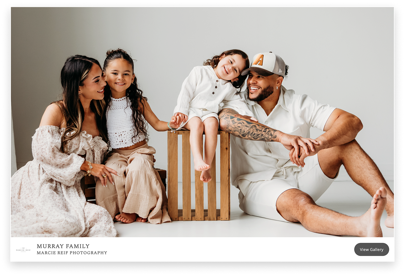A smiling family of four, dressed in coordinated white and beige outfits, poses together in a bright studio setting, with the father sitting on the floor, the mother seated next to their older daughter, and their youngest child sitting on a wooden crate in the center.