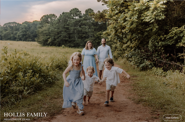 A family of five is walking on a dirt path in a field, with the three children running ahead holding hands. The parents are walking behind them, holding hands as well, with trees and a field in the background.