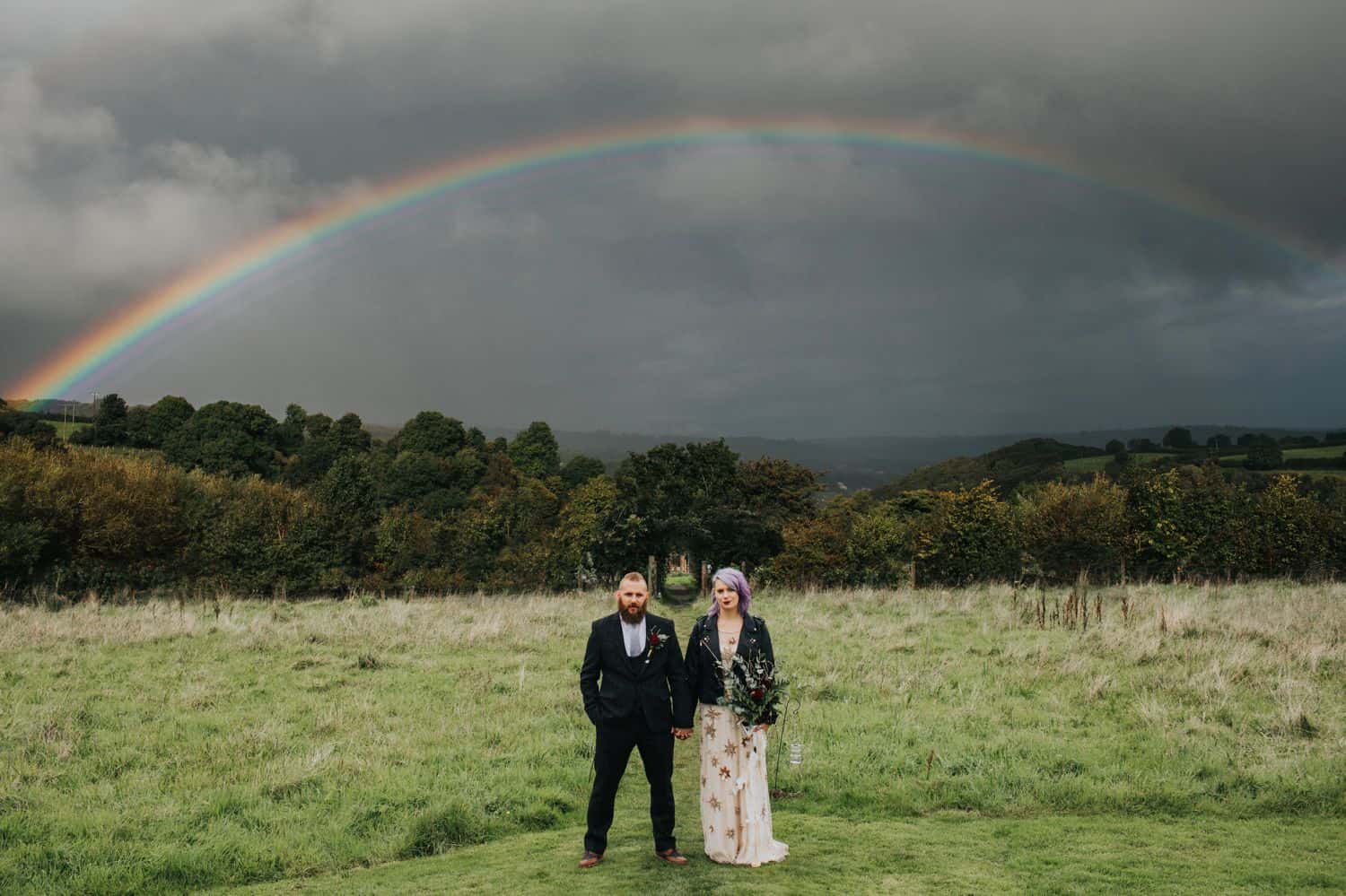How To Make A Photography Website Your Dream Clients Can’t Resist: A punk-rock bride and groom stand in a field below a rainbow glowing through ominous clouds.