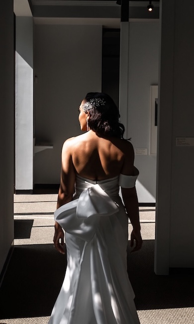 A woman in a white strapless gown with a large bow in the back walks down a hallway, with light streaming in from the left. She is wearing a jeweled headpiece and long earrings.