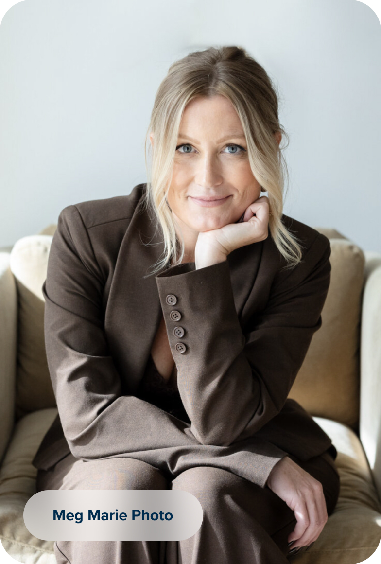 A blonde woman with blue eyes sits in a chair, wearing a brown suit and resting her chin on her hand. She is looking directly at the camera with a slight smile.