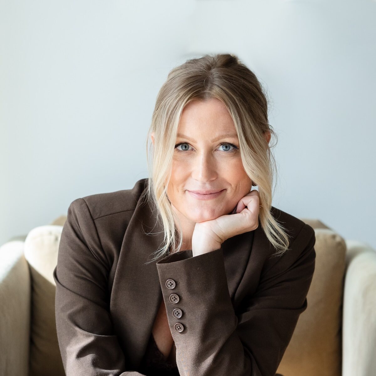 A blonde woman with blue eyes sits in a beige chair, wearing a brown suit and resting her chin on her hand while looking at the camera. The background is a plain white wall.