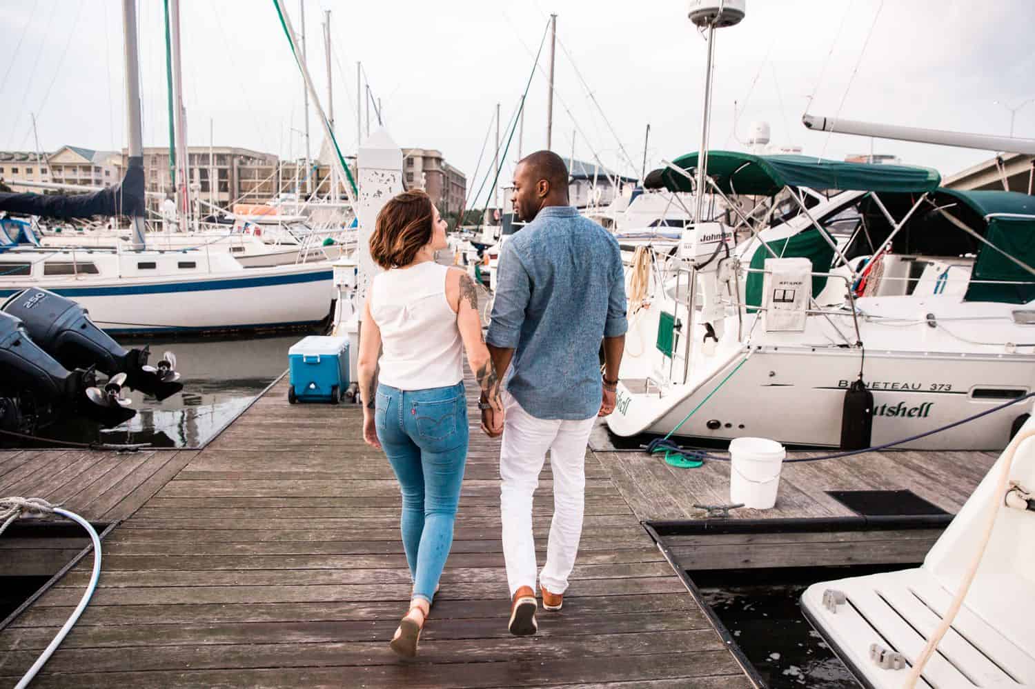 A man and woman walk along the dock of the bay with yachts and sailboats in the background.