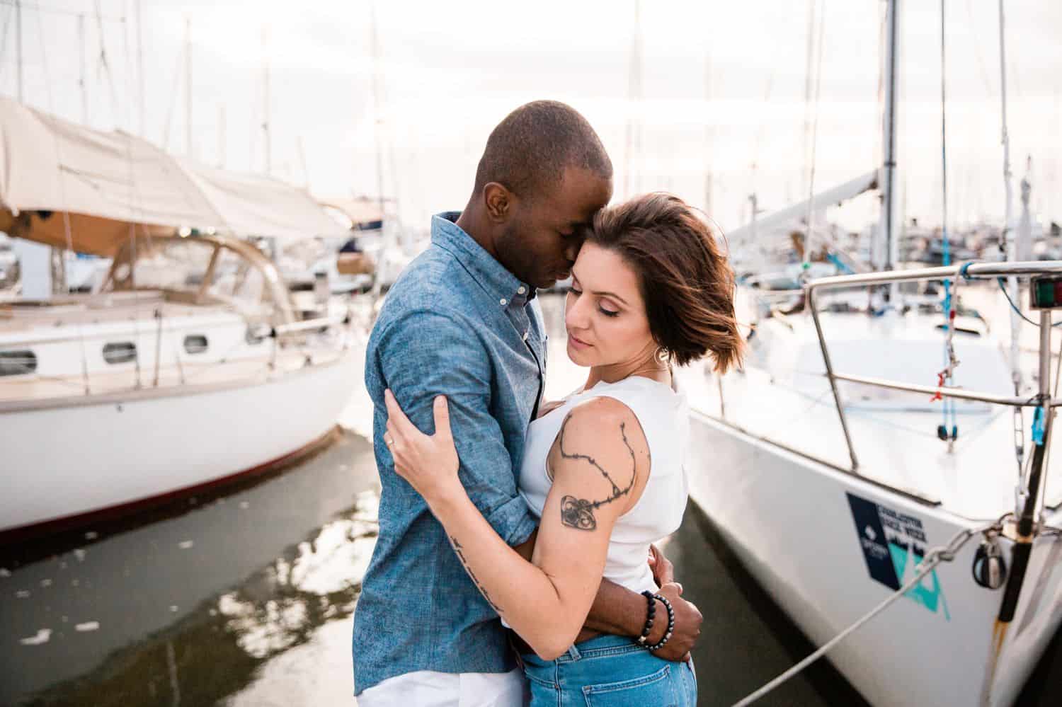 A Black man in a denim shirts and a White woman in a white shirt hold each other close on the dock of the bay