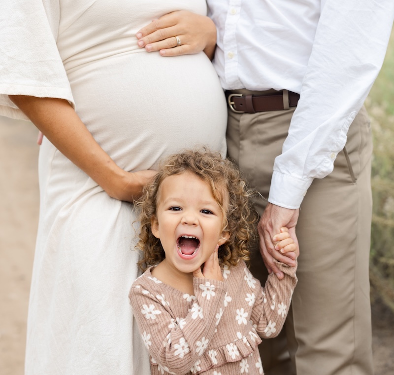 Happy toddler holds parents hands; pregnant mothers belly visible