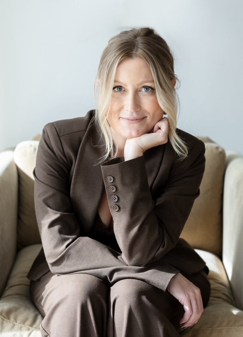 A blonde woman with blue eyes sits in a beige armchair, wearing a brown pantsuit and resting her chin on her hand while smiling at the camera. The jacket has four buttons on the sleeve.