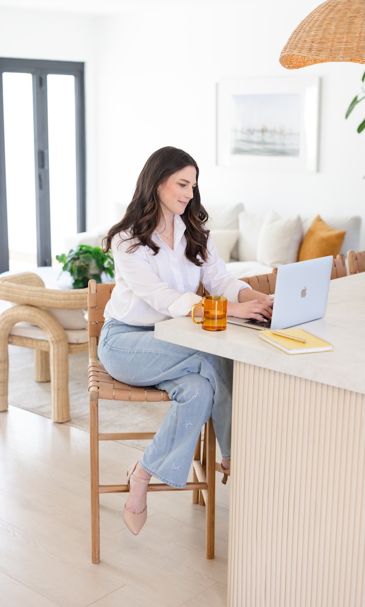 Woman in white shirt and jeans works on laptop at kitchen counter
