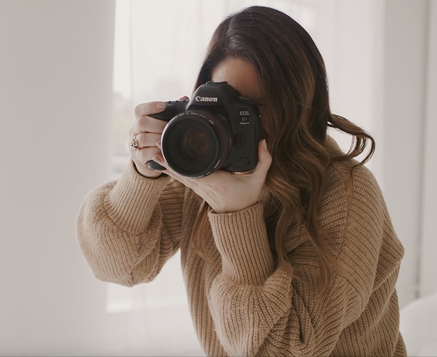 A woman with long brown hair wearing a tan sweater holds a Canon EOS 5D Mark IV camera up to her face, ready to take a picture. The background is a soft, neutral tone.